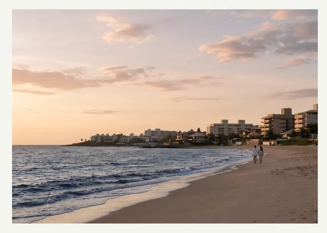 Paisagem costeira ao entardecer com mar calmo, ondas na areia e casas à beira-mar, com duas pessoas caminhando na praia
